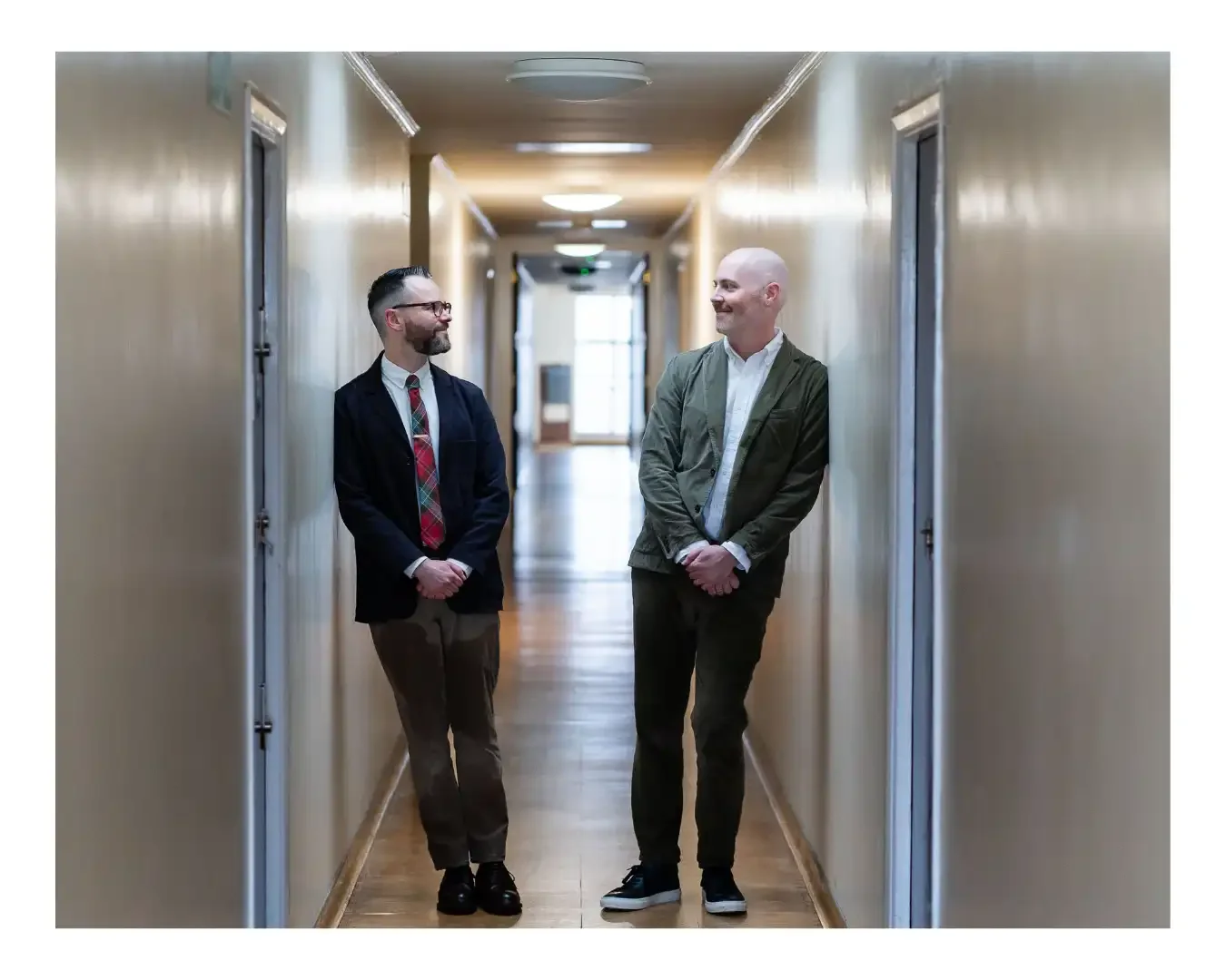 gay wedding couple standing in corridor leaning against opposite walls smiling at each other