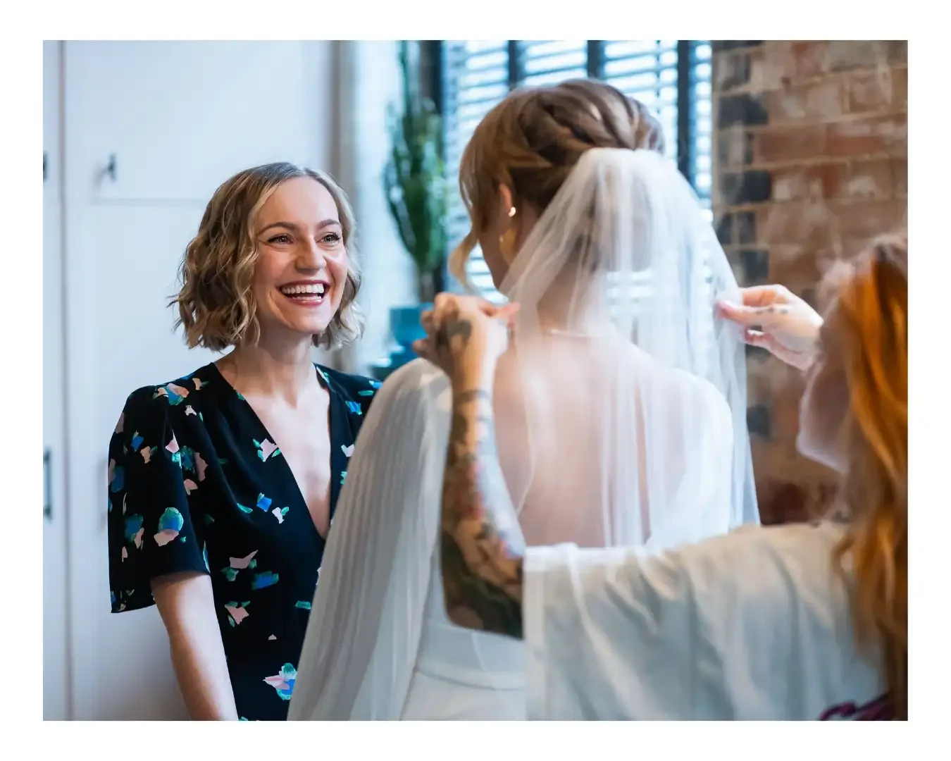 Bridesmaid smiling at bride while bride is having her veil put on by stylist