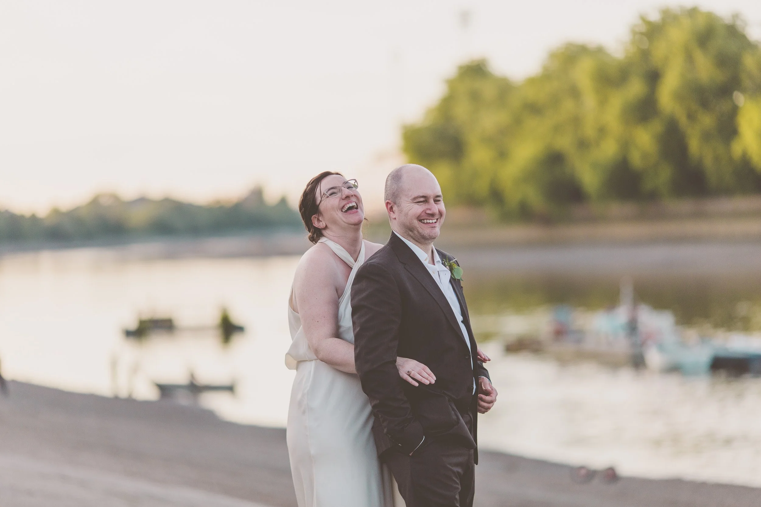 Bride holding groom around waist as both laugh in sunset picture on  Putney Embankment with River Thames in background