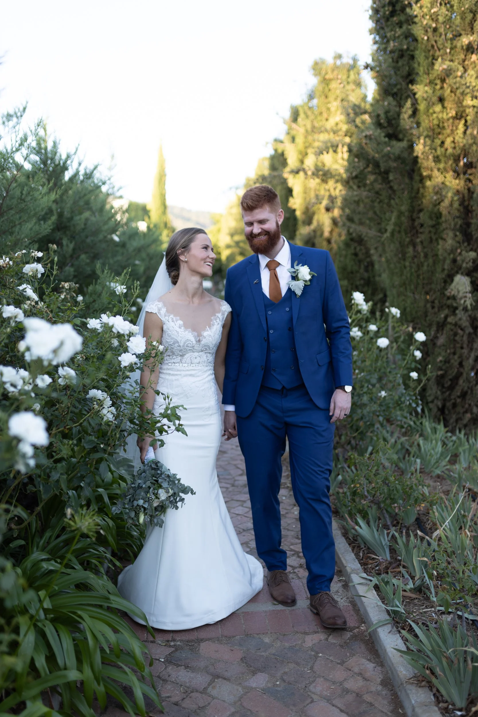 Photo of a woman wearing a white wedding dress and a man wearing a blue wedding suit holding hands and walking down a path surrounded by greenery and flowers edited with Archipelago Lightroom Presets