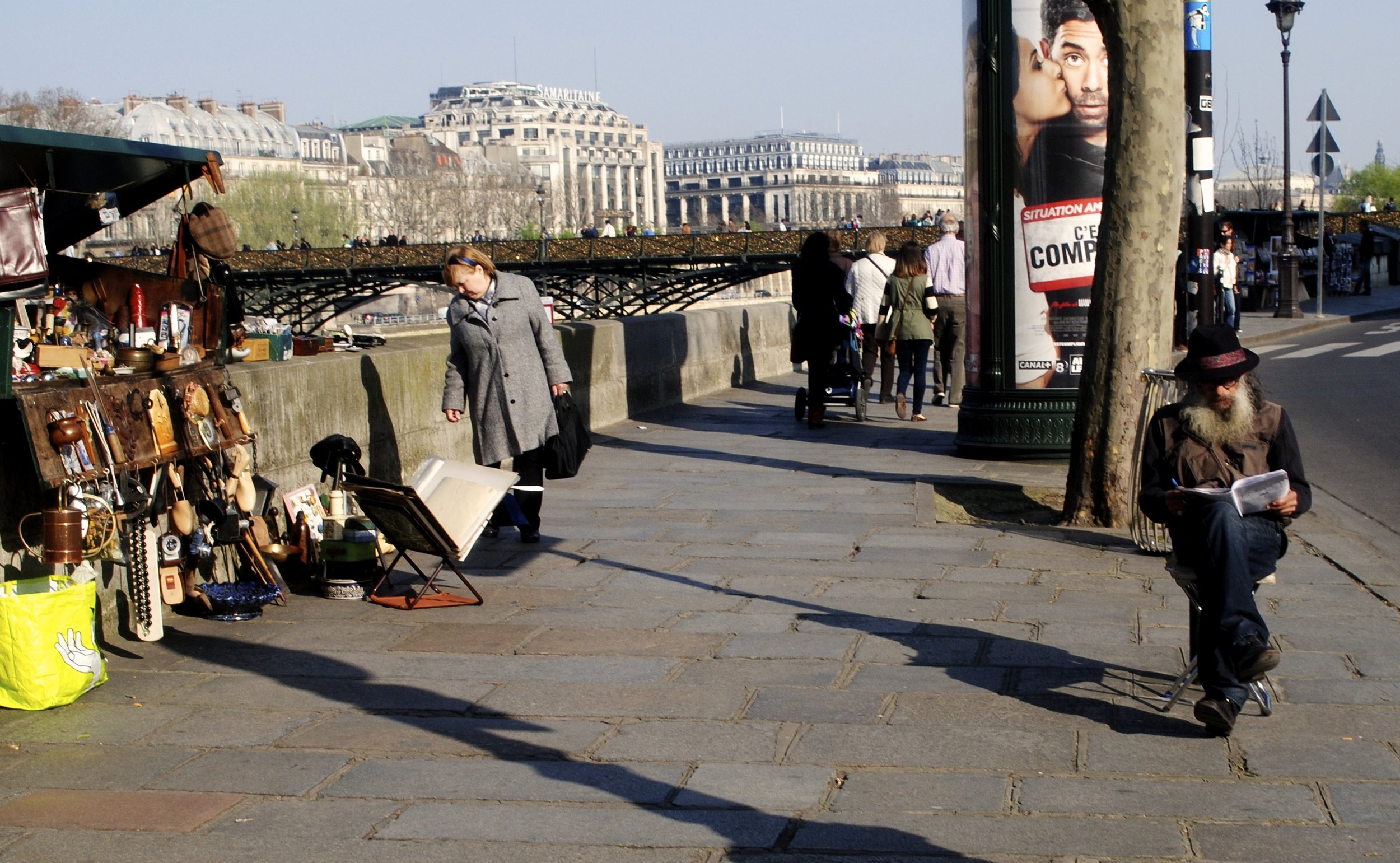 A woman looks at the market of items as she walks by. Meanwhile, the market owner reads to himself on the sidewalk. // Paris, France