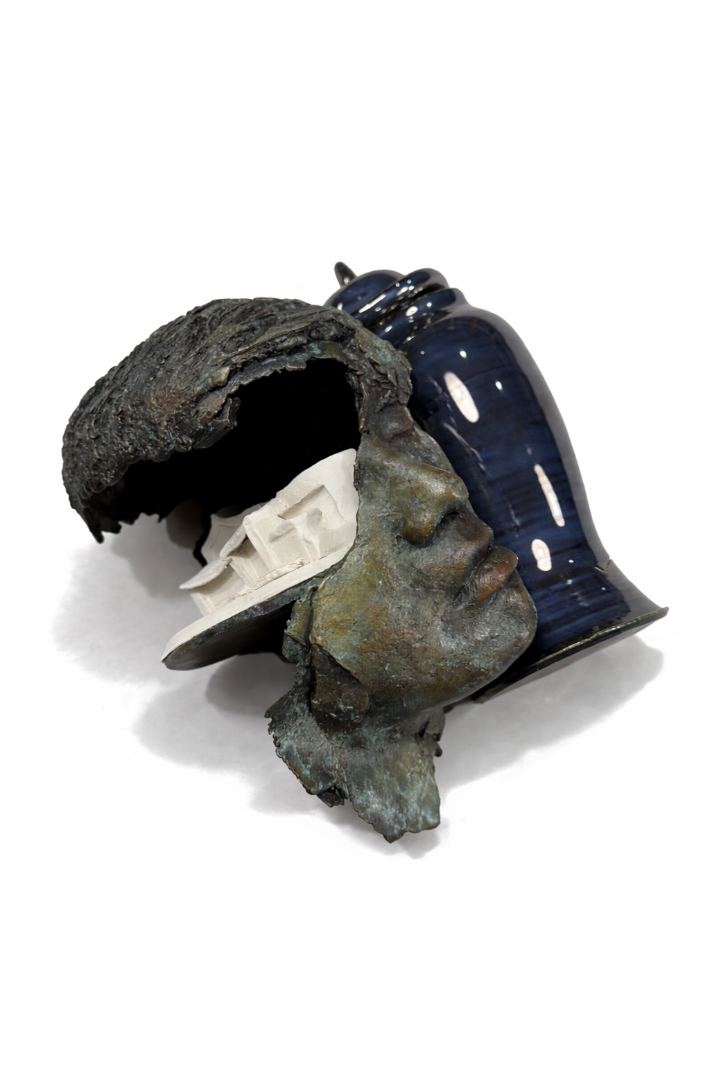 A mixed media sculpture combines a human face with a dark hat, a model building, and a ceramic vessel, creating an abstract artistic composition.