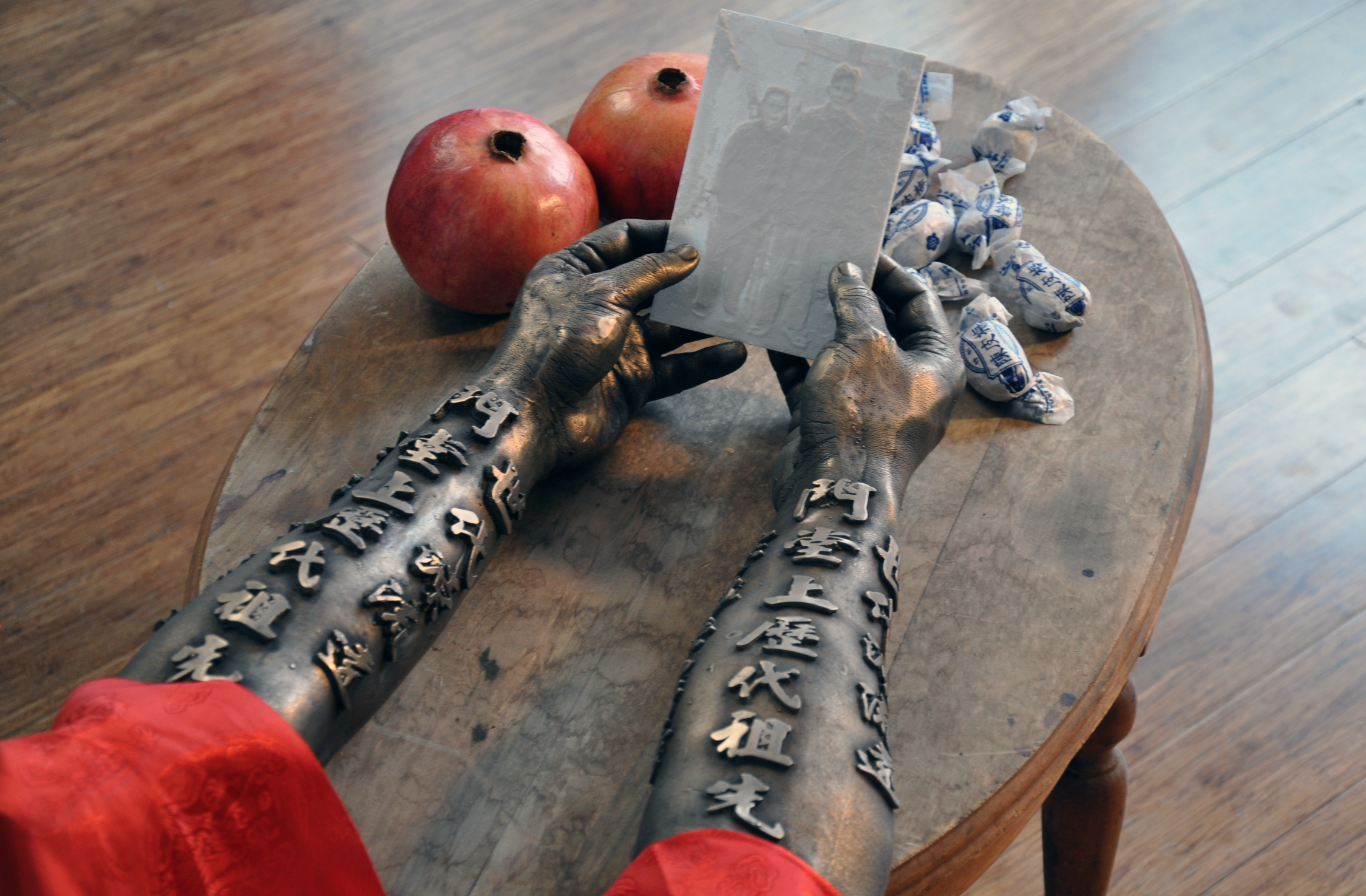Bronze-colored tattooed arms with Chinese characters, holding a piece of paper, sitting at a wooden table with two pomegranates and wrapped candies on a round wooden table.