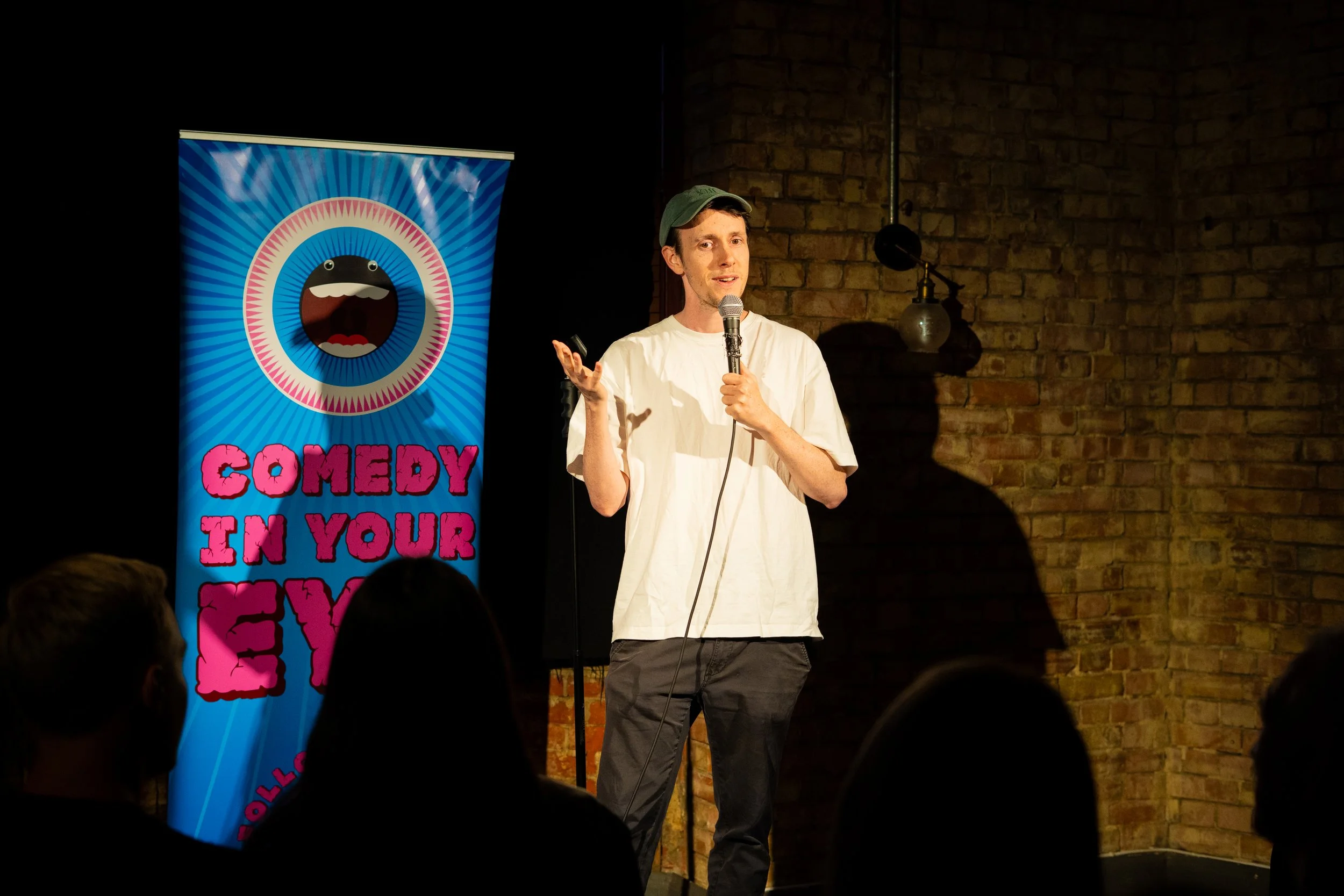 A man wearing a green cap and white t-shirt is performing stand-up comedy on stage at a comedy club. Behind him, there is a colorful poster with a cartoonish eyes and mouth character and pink text saying 'Comedy in your'.