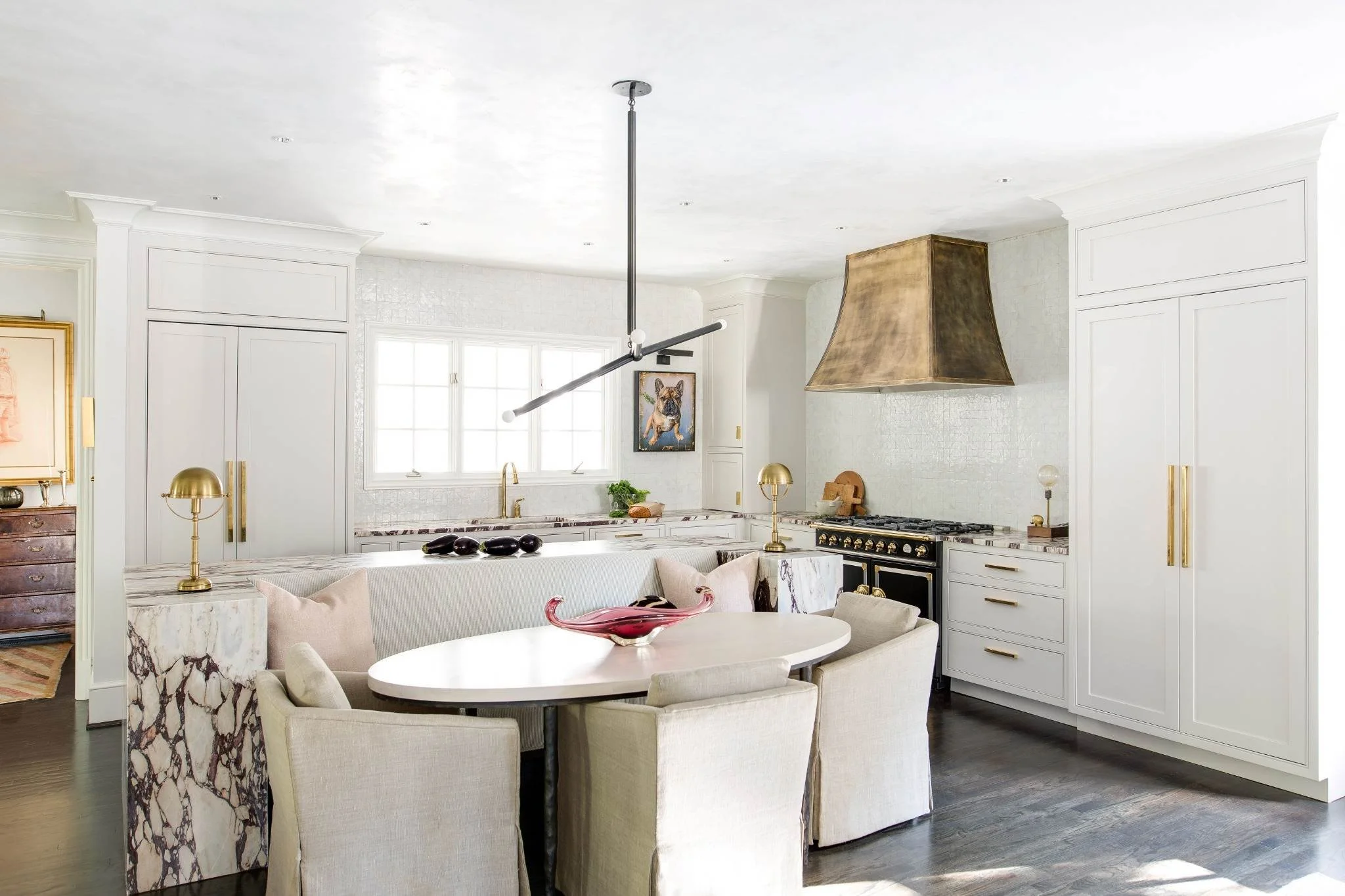Bright luxury kitchen with white cabinetry, dramatic marble countertops, brass accents, a statement range hood, and a built-in dining area with upholstered seating.