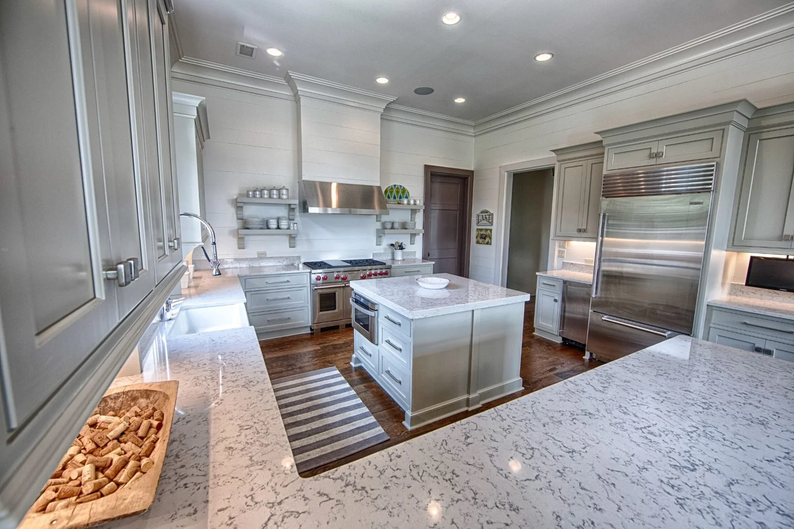 Light gray custom kitchen cabinetry with white stone countertops, a center island, stainless steel appliances, and warm wood flooring.