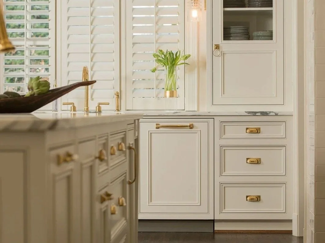 White inset kitchen cabinetry with brass hardware, integrated drawers, glass-front storage, and a sink beneath shuttered windows.
