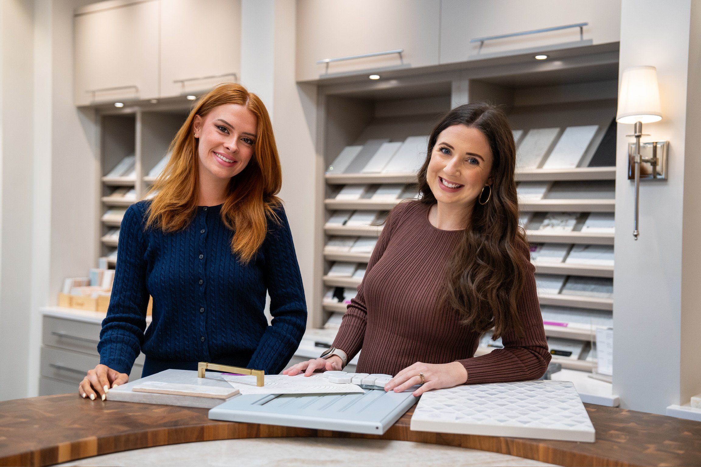 Two kitchen designers standing in a showroom reviewing cabinet door styles and material samples, surrounded by organized display racks of finishes and surfaces.