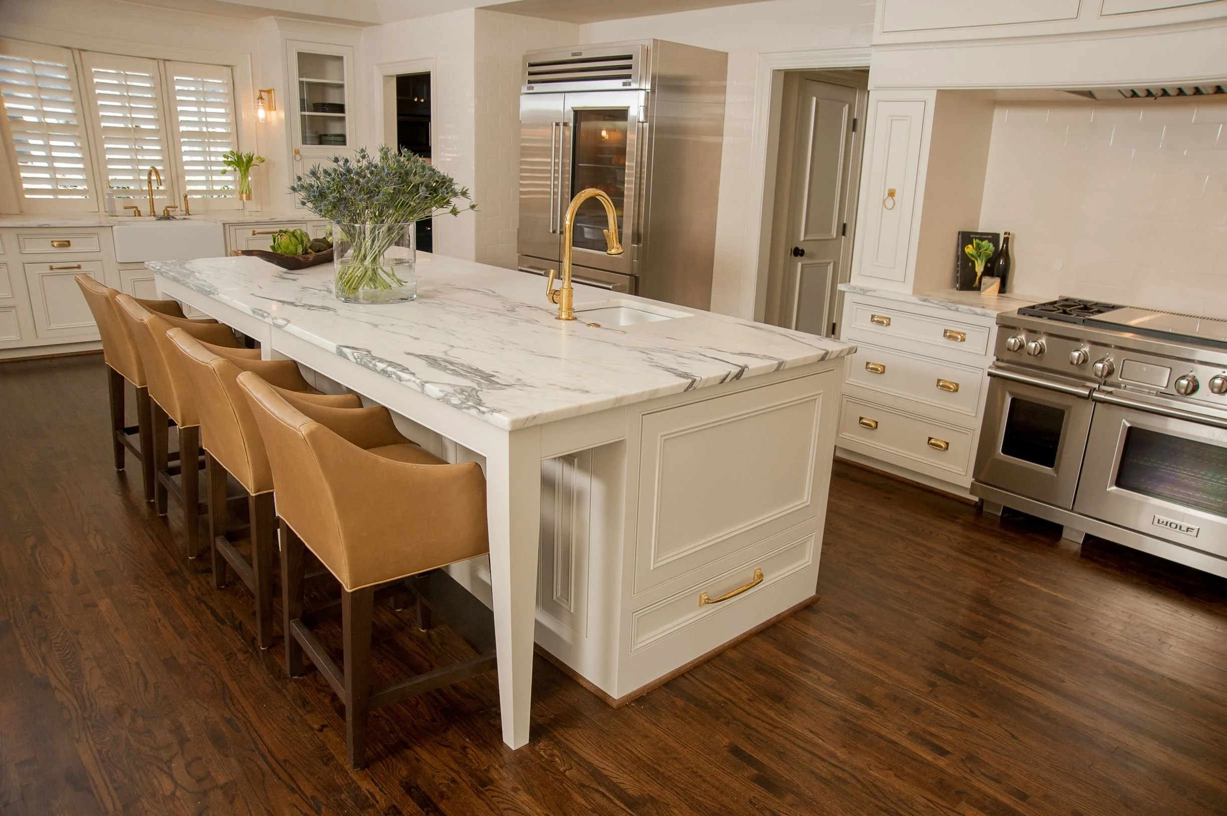 Luxury kitchen in Mountain Brook, Alabama featuring custom white cabinetry, oversized marble waterfall island, brass faucet and hardware, leather bar stools, and Wolf appliances.
