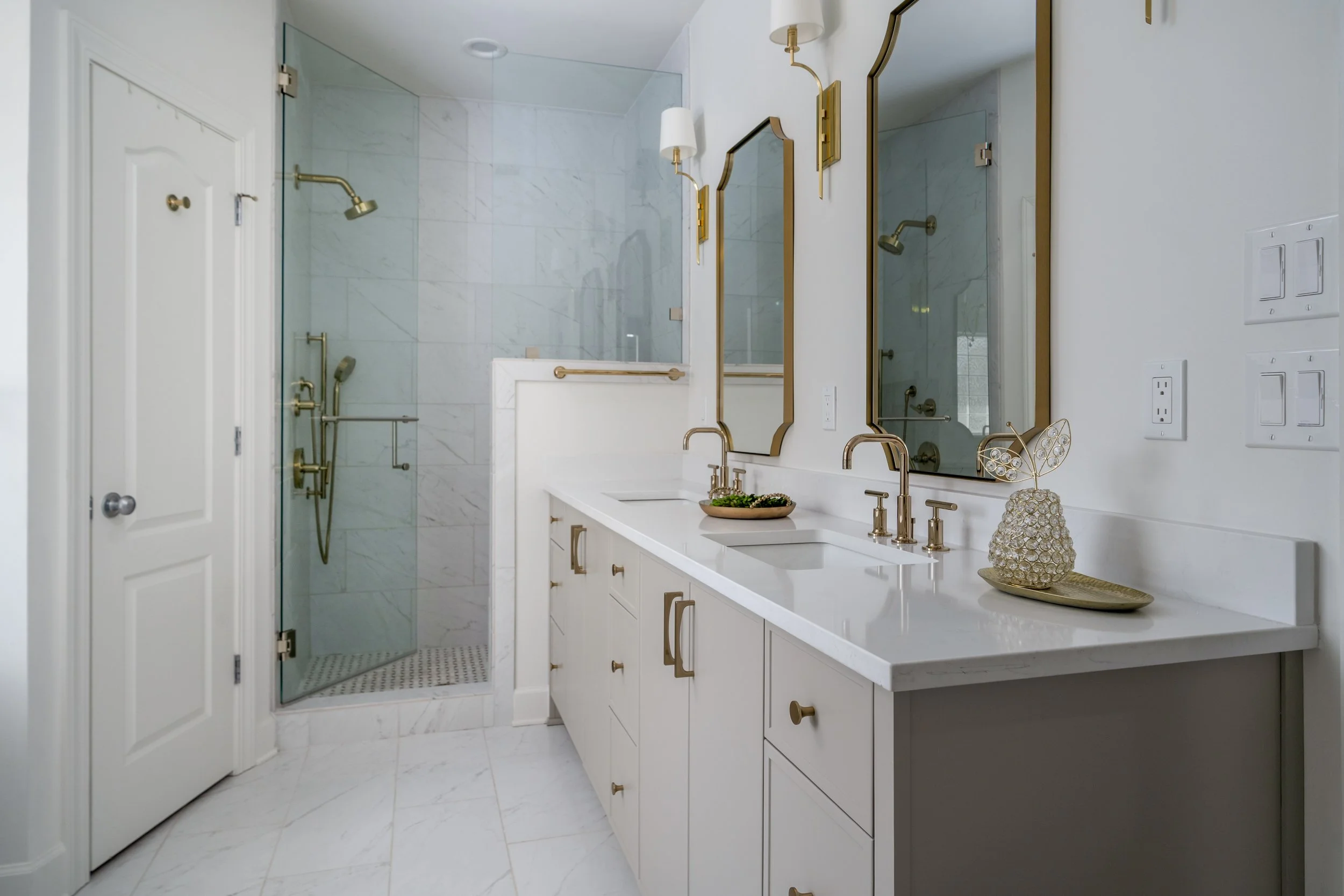Modern white bathroom with a double vanity, brass fixtures, arched mirrors, and a glass-enclosed walk-in shower.
