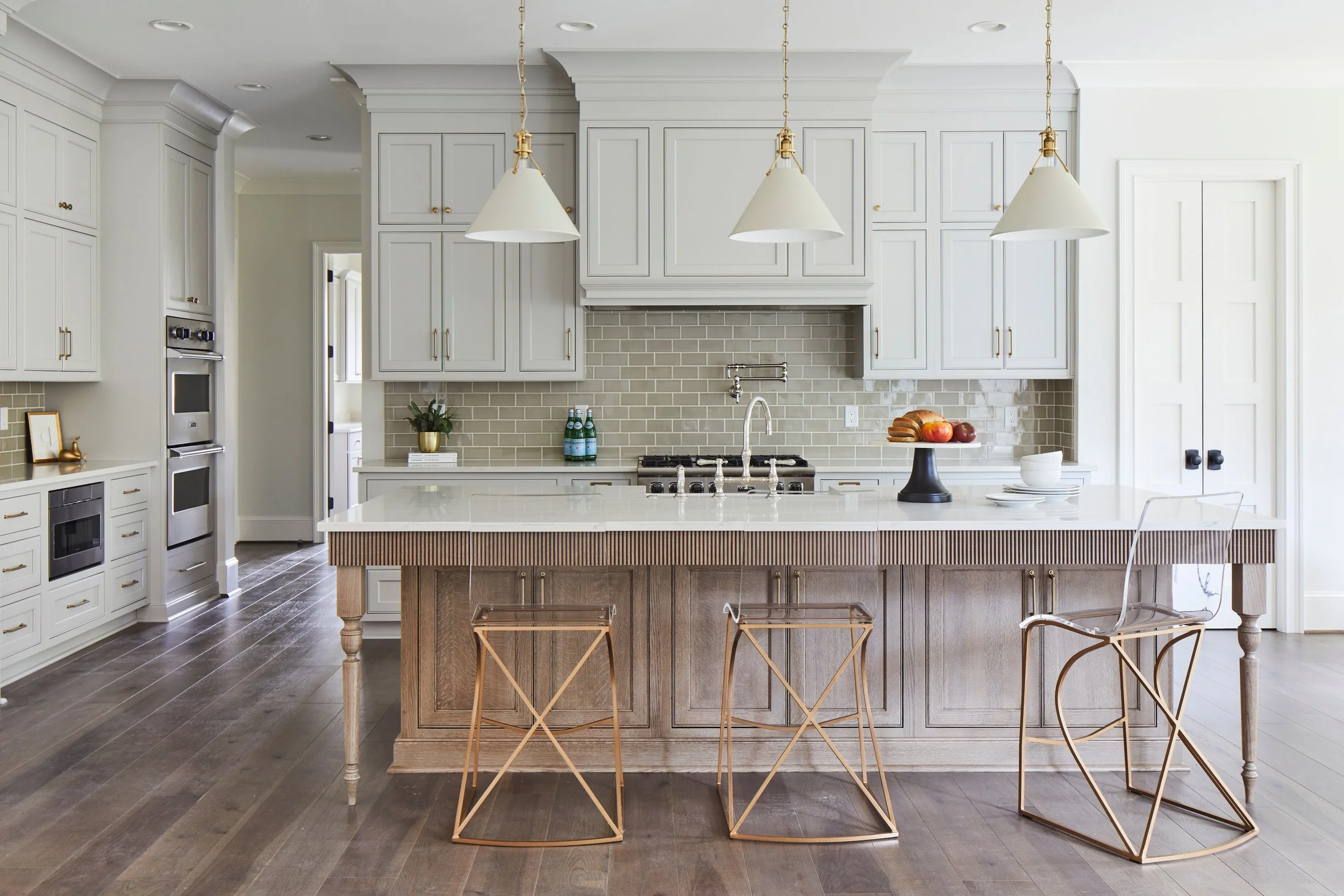Custom luxury kitchen featuring white inset cabinetry, a stained wood island with reeded detailing and furniture-style legs, quartz countertops, brass hardware, pendant lighting, and a soft gray subway tile backsplash in a Tuscaloosa home.