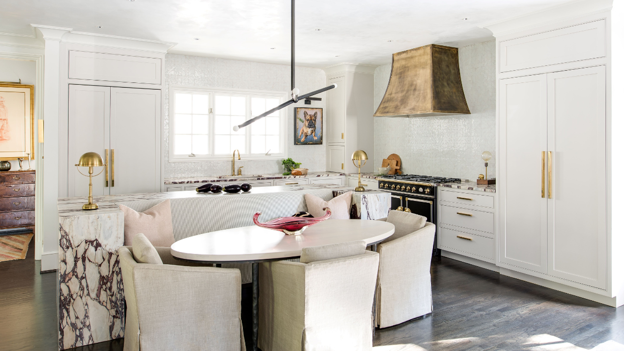 Bright white kitchen featuring custom cabinetry, a marble waterfall island, brass hardware, a statement range hood, and a built-in dining nook with upholstered seating.