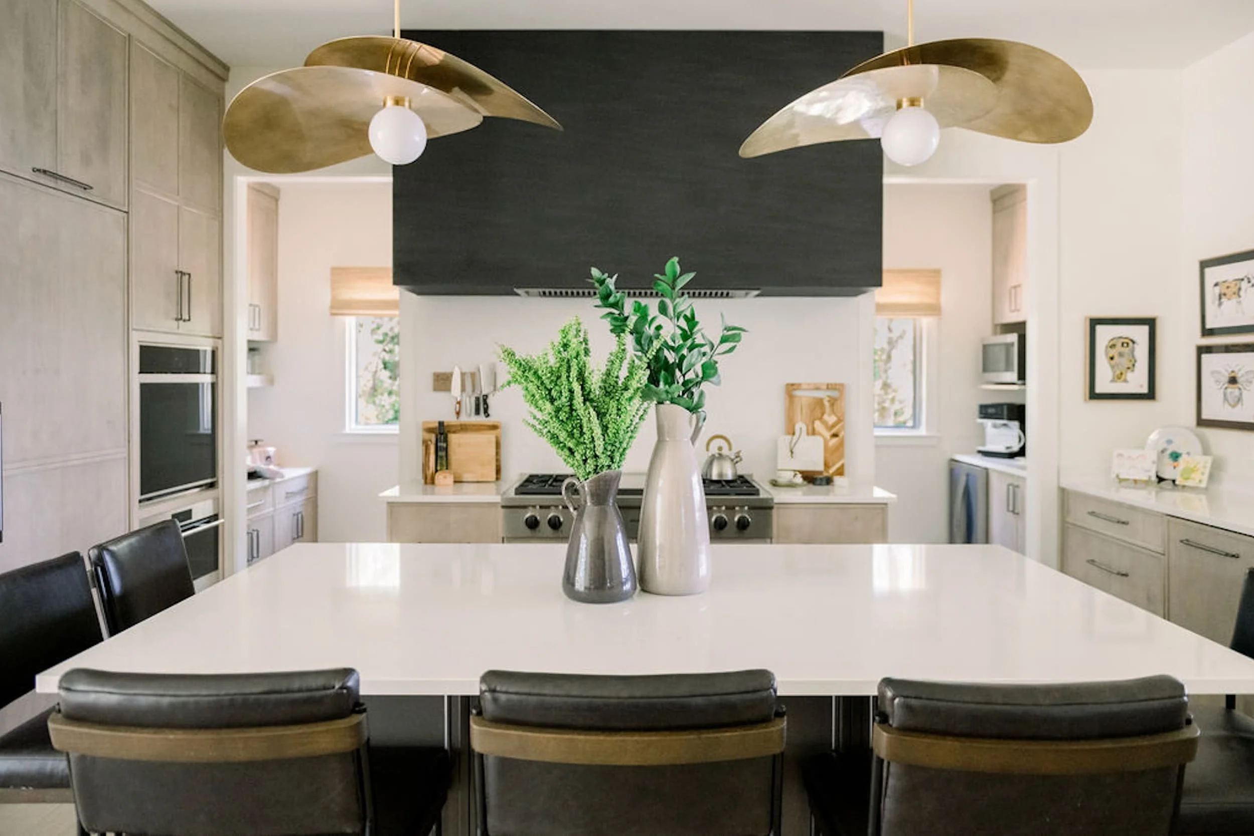 Modern luxury kitchen featuring a large white island, warm wood cabinetry, black range hood, sculptural brass pendant lighting, and contemporary finishes in a custom Alabama home.