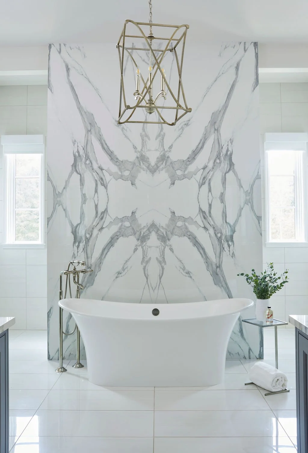 Freestanding white soaking tub positioned in front of a dramatic bookmatched marble slab accent wall with gray veining, complemented by a brass floor-mounted tub filler and brass lantern-style pendant lighting in a luxury bathroom.