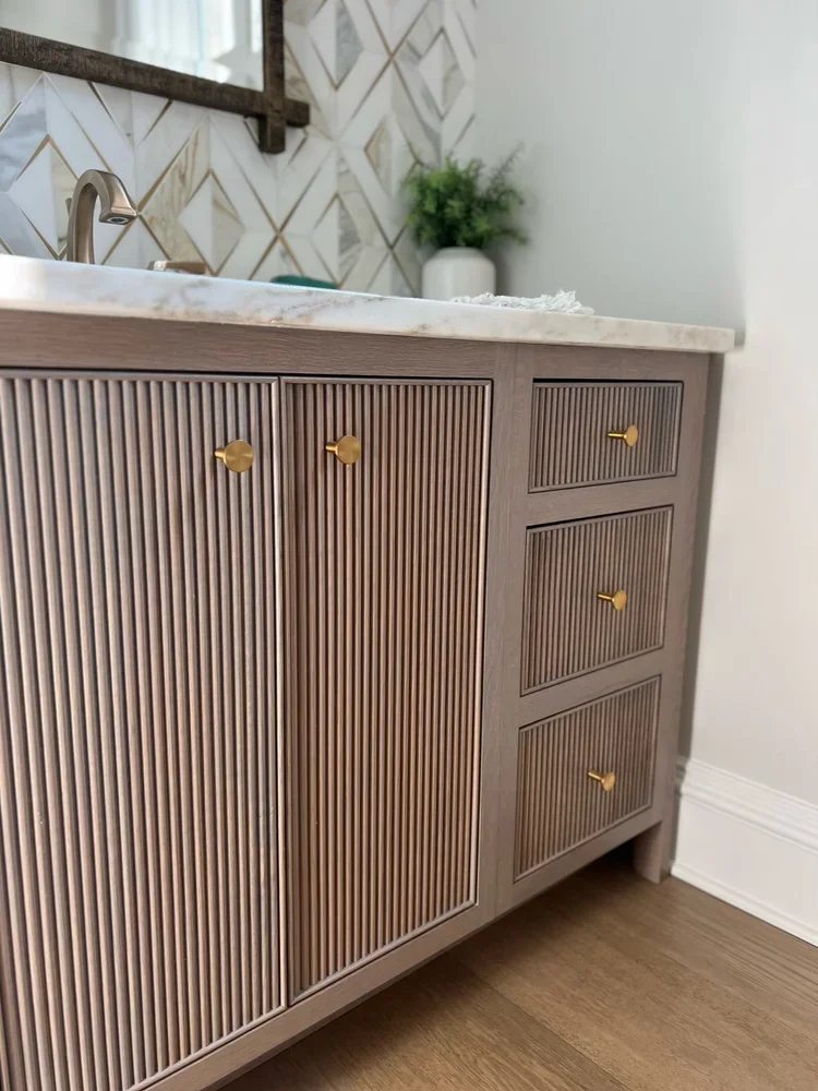 Custom bathroom vanity featuring reeded cabinet doors and drawer fronts in a warm wood finish, topped with a marble countertop and accented with round brass knobs for a refined, architectural look.