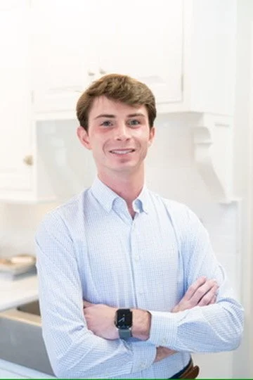 A young man with light skin and brown hair smiling while standing with arms crossed in front of a light-colored wall decorated with blue botanical illustrations.