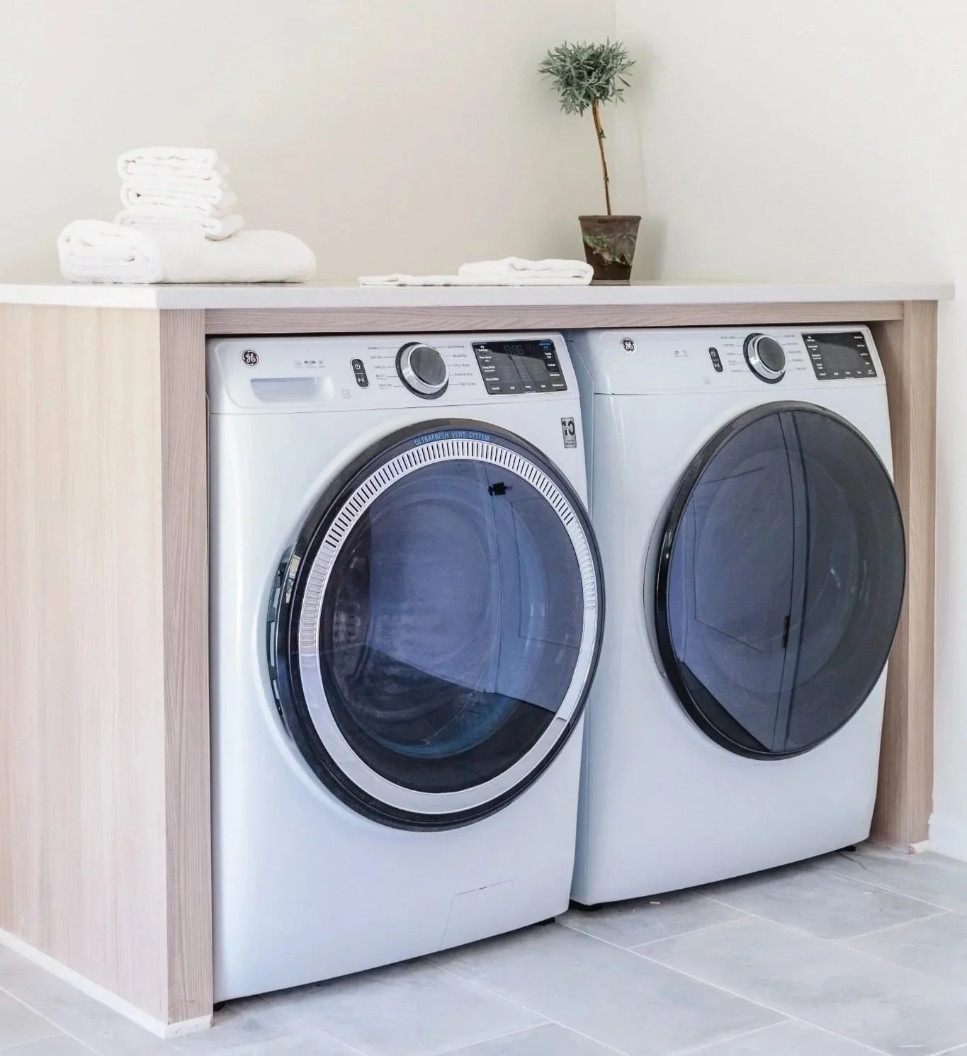Laundry room with side-by-side front-loading washer and dryer beneath a built-in folding counter with light wood surround.