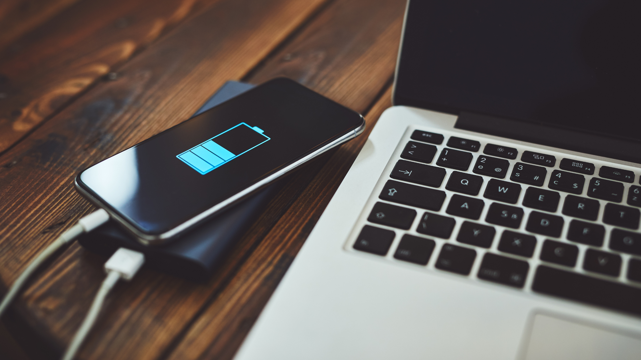 Smartphone charging from a portable power bank beside a laptop on a wooden table, showing a full battery icon for backup power during a power outage.