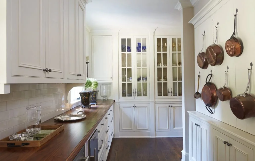 White custom cabinetry, glass-front display storage, wood countertops, and hanging copper cookware create a functional butler’s pantry.