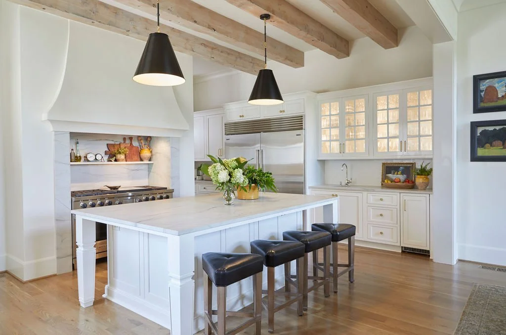 Bright kitchen with exposed wood ceiling beams and a large island featuring a thick built-up countertop with an eased edge, paired with white cabinetry, black pendant lighting, and a custom range hood.