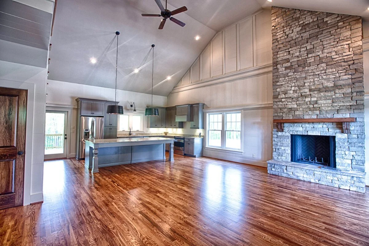 Vaulted open kitchen and living room with a stone fireplace and wood floors in an Alabama home.