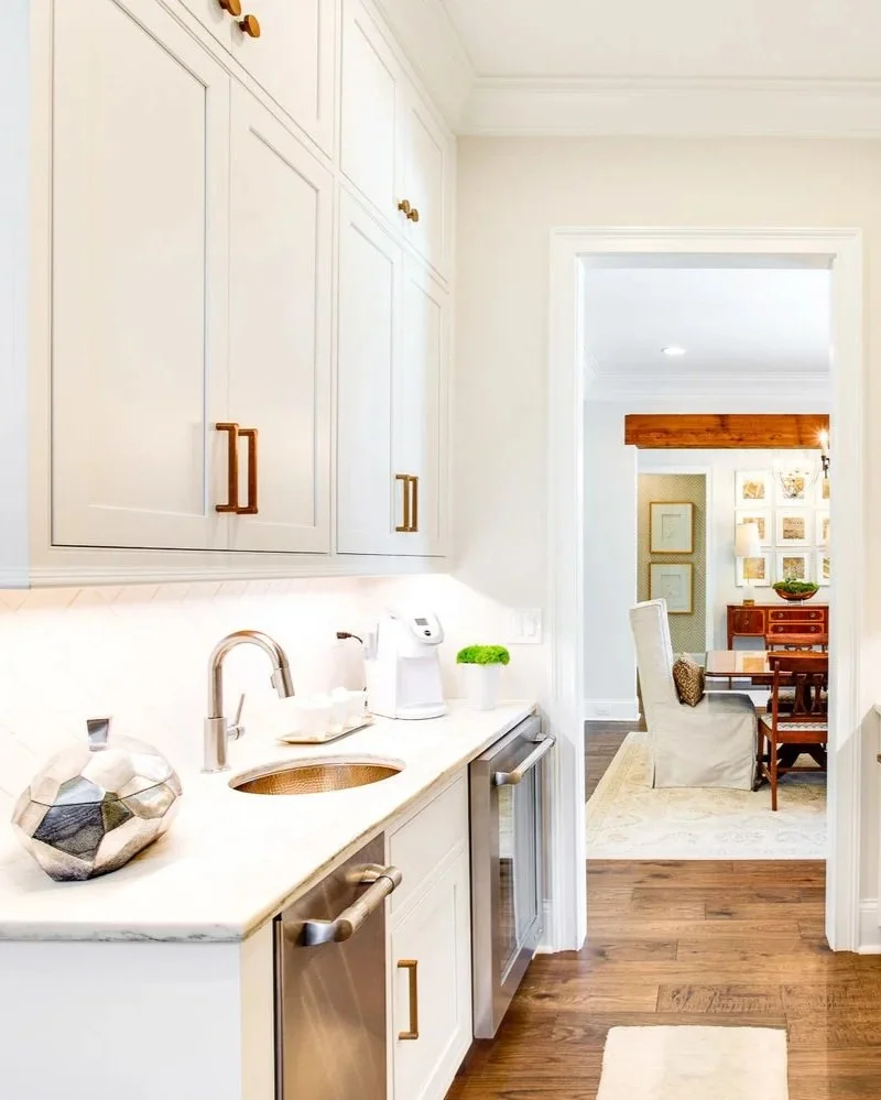 Custom kitchen beverage station with white inset cabinetry, brass hardware, under-cabinet lighting, a prep sink, beverage refrigerator, and quartz countertops connecting the kitchen and dining room in a luxury home.