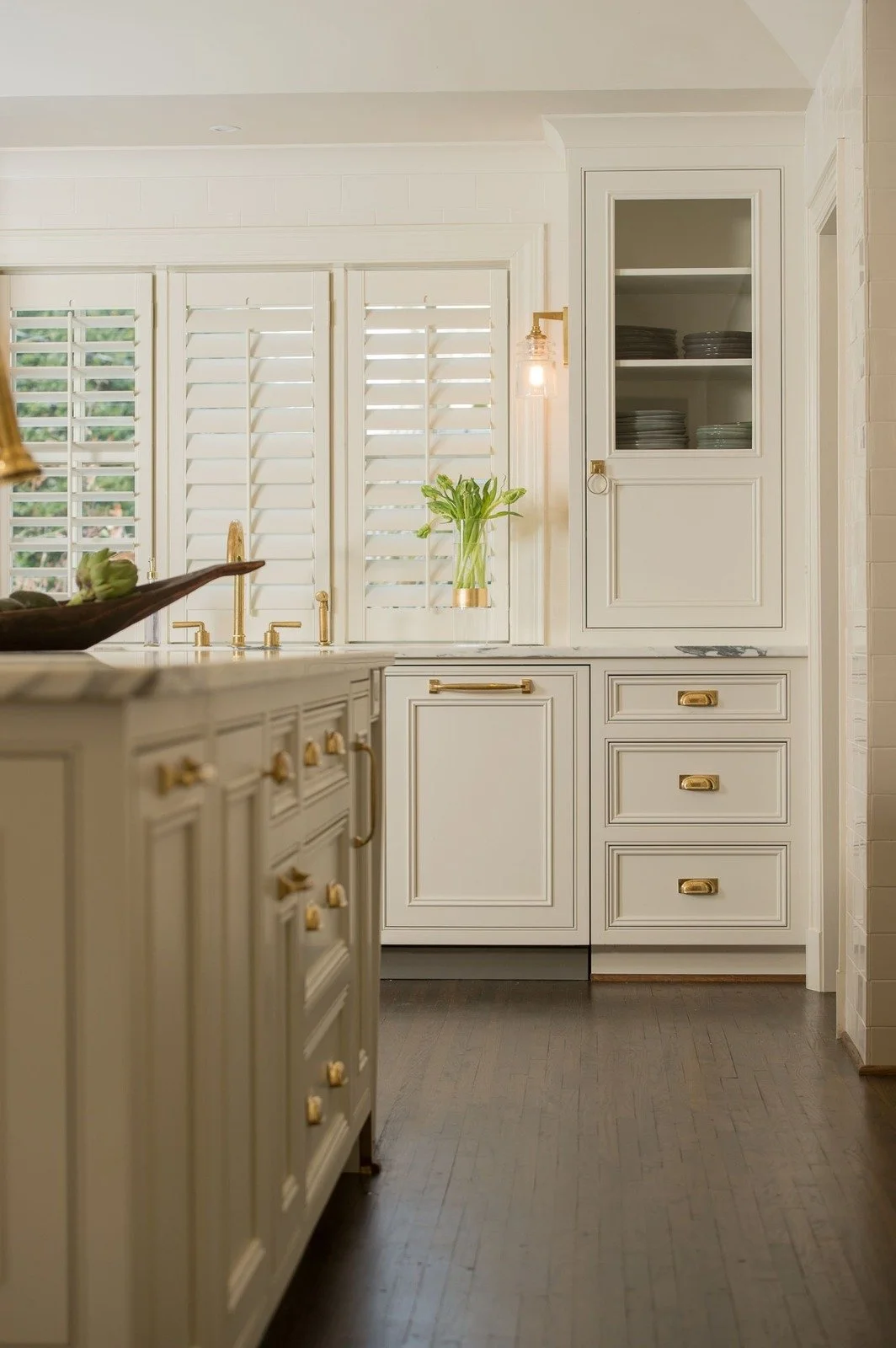 White custom cabinetry, glass-front storage, brass hardware, and a bright sink area create a polished and functional kitchen wall.