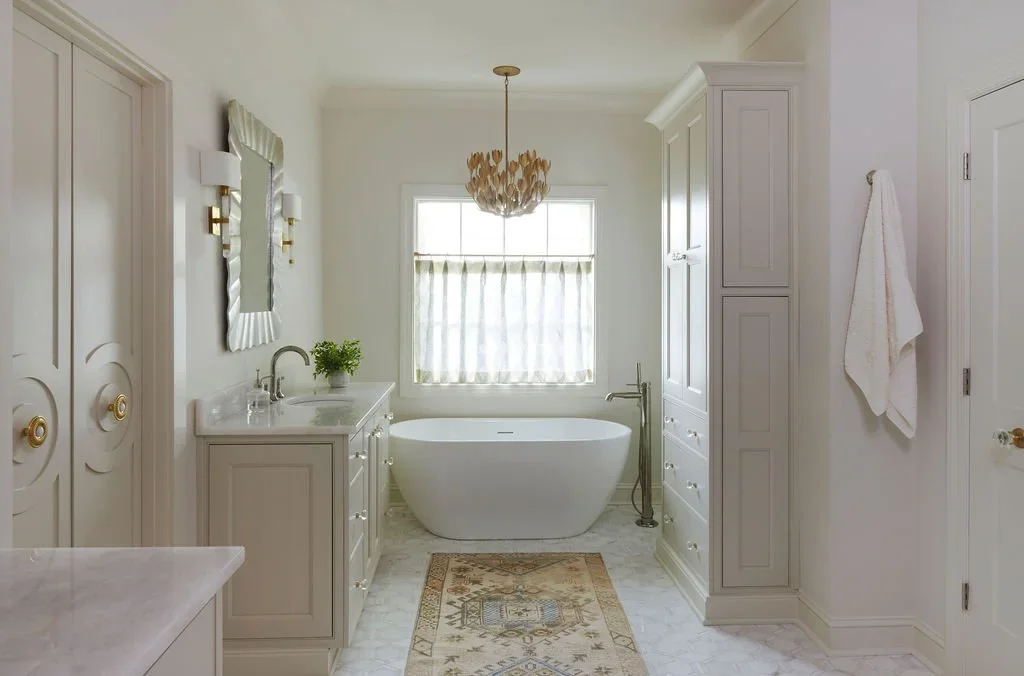 Elegant white primary bathroom featuring custom inset cabinetry, a freestanding soaking tub beneath a large window, marble flooring, brass accents, tall storage cabinets, and a decorative statement chandelier.
