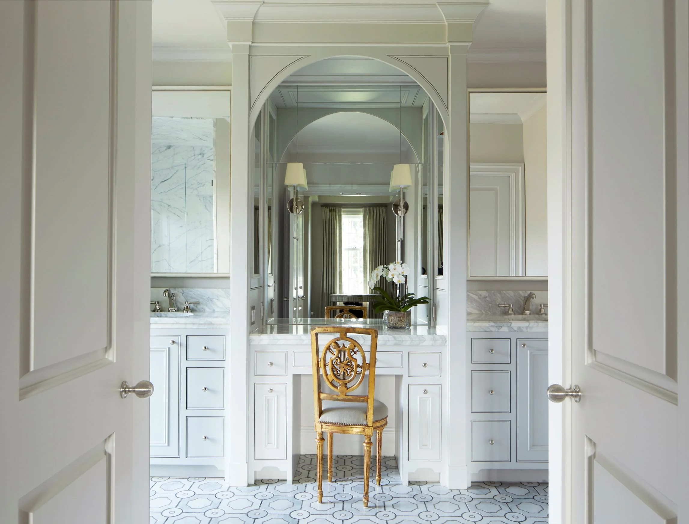 Elegant bathroom vanity featuring custom light gray cabinetry, marble countertops, mirrored architectural detailing, hexagon tile flooring, and a gold accent chair in a luxury Alabama home.