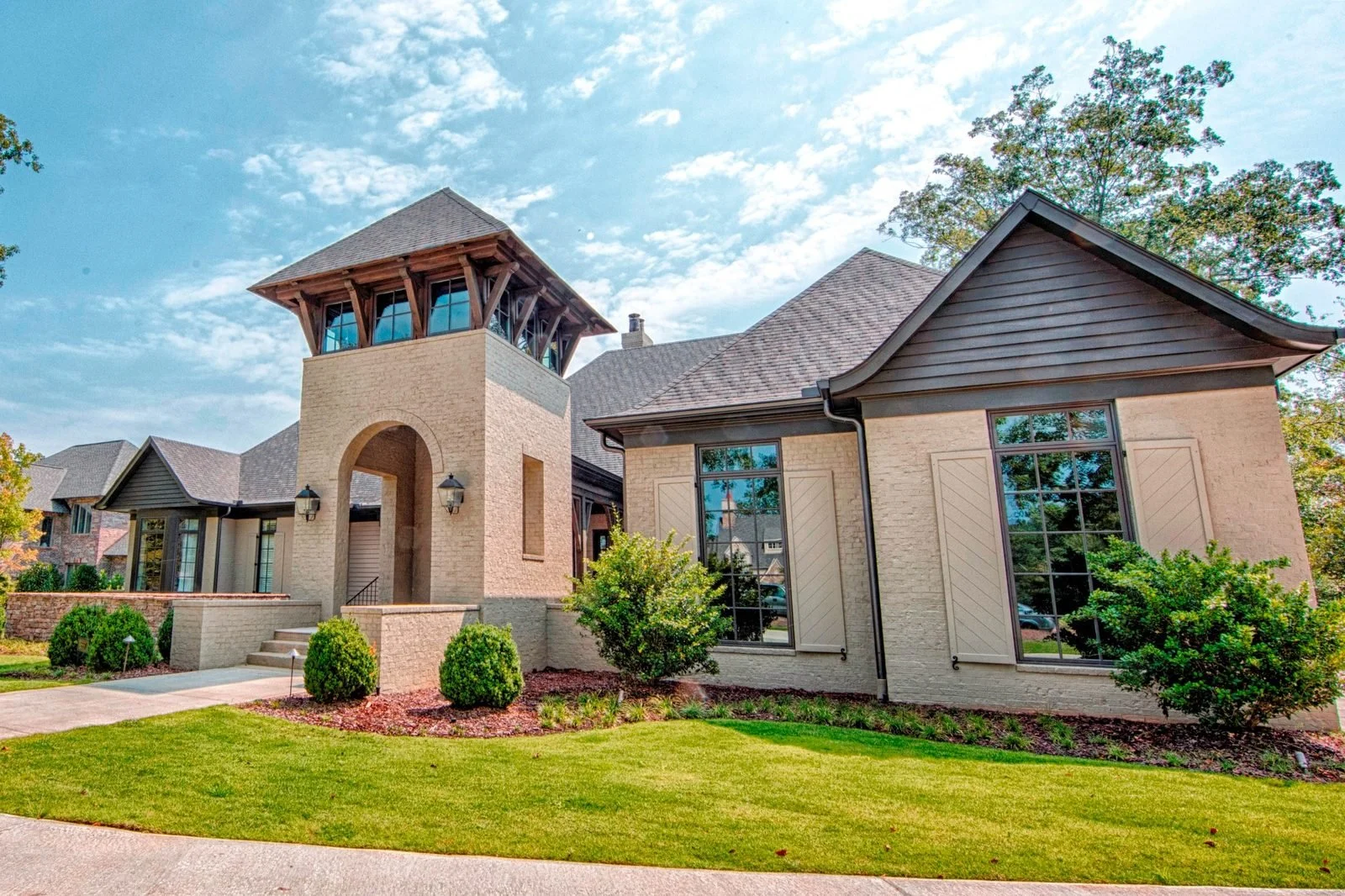 Custom luxury home exterior in Tuscaloosa, Alabama featuring painted brick, a dramatic arched tower entrance with exposed timber details, oversized black-framed windows, decorative shutters, and professionally landscaped grounds.