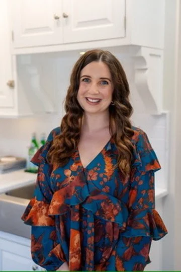 A woman with curly brown hair, blue eyes, and a white sleeveless blouse posing in front of a blue and gray abstract floral background.