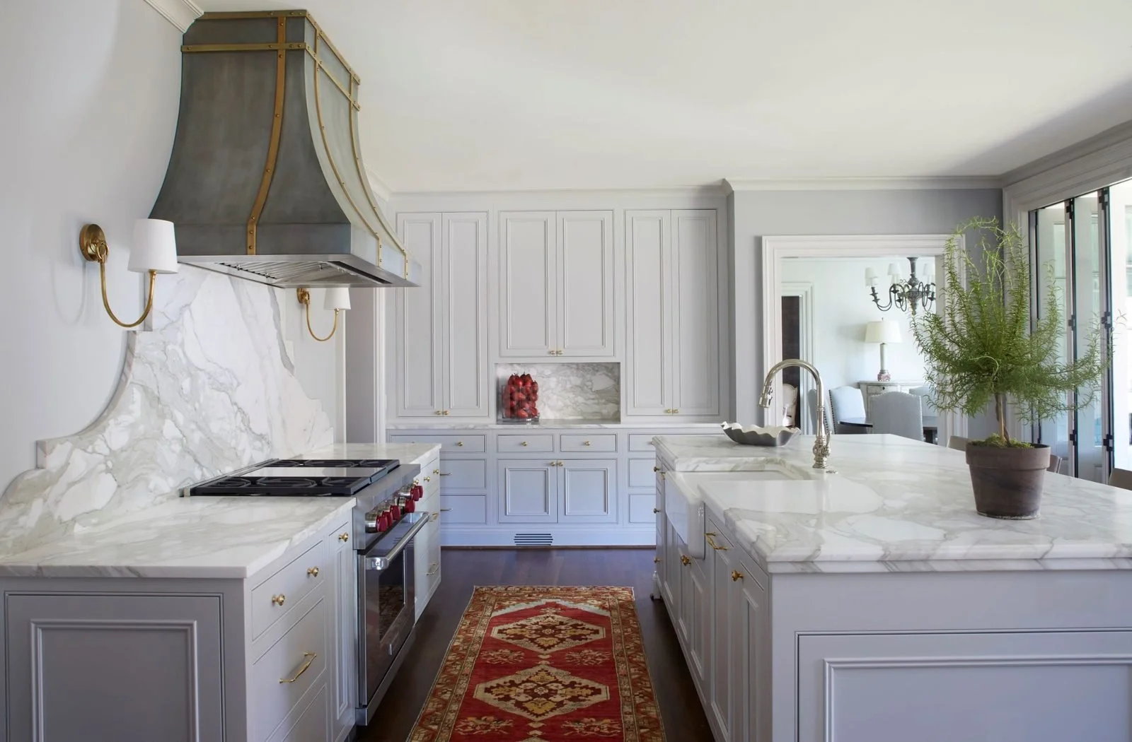Custom kitchen with light painted cabinetry, marble countertops and backsplash, large island, and statement brass range hood.
