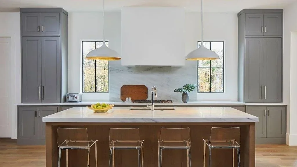 Modern kitchen with gray cabinetry, a white plaster range hood, marble backsplash, wood island, and acrylic bar stools.