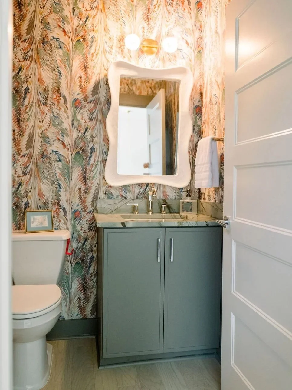Bathroom vanity with Taiga green cabinetry, marble countertop, and patterned wallpaper, showcasing a nature-inspired cabinet color trend designed by Toulmin Kitchen & Bath in Tuscaloosa, Alabama.