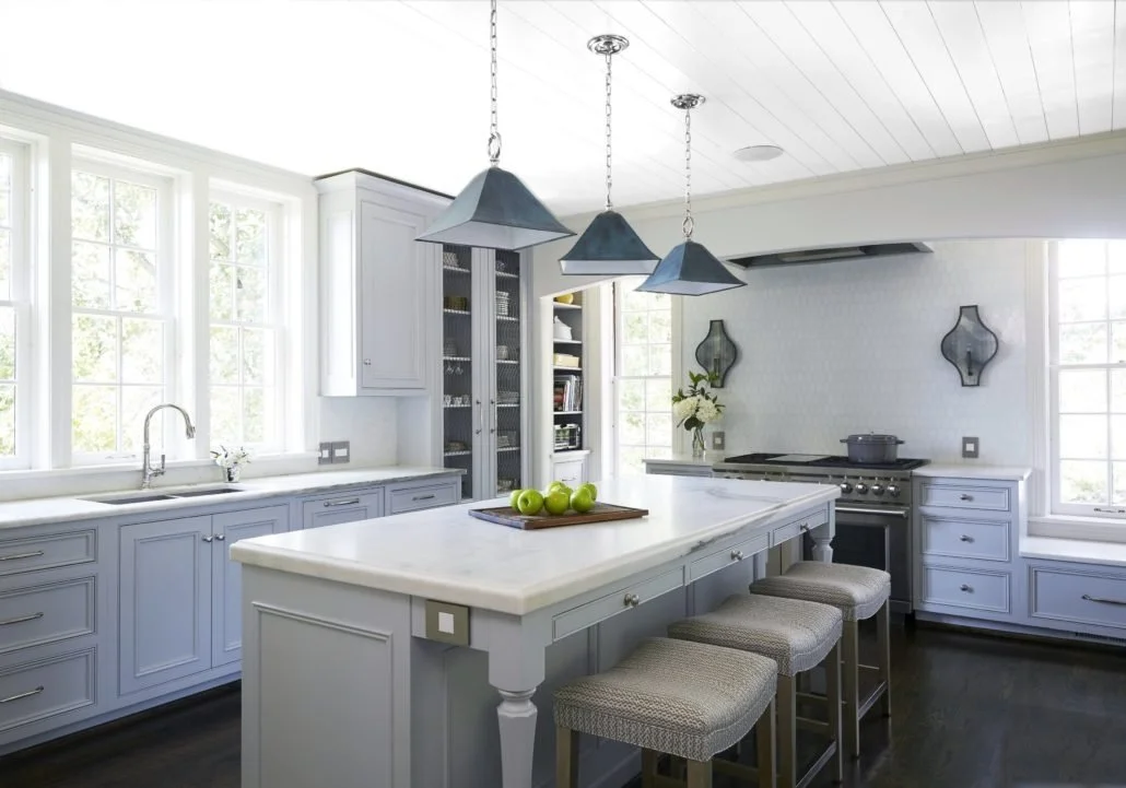 Bright kitchen with soft gray cabinetry, a large white island with upholstered stools, dark pendant lights, and abundant natural light from oversized windows.