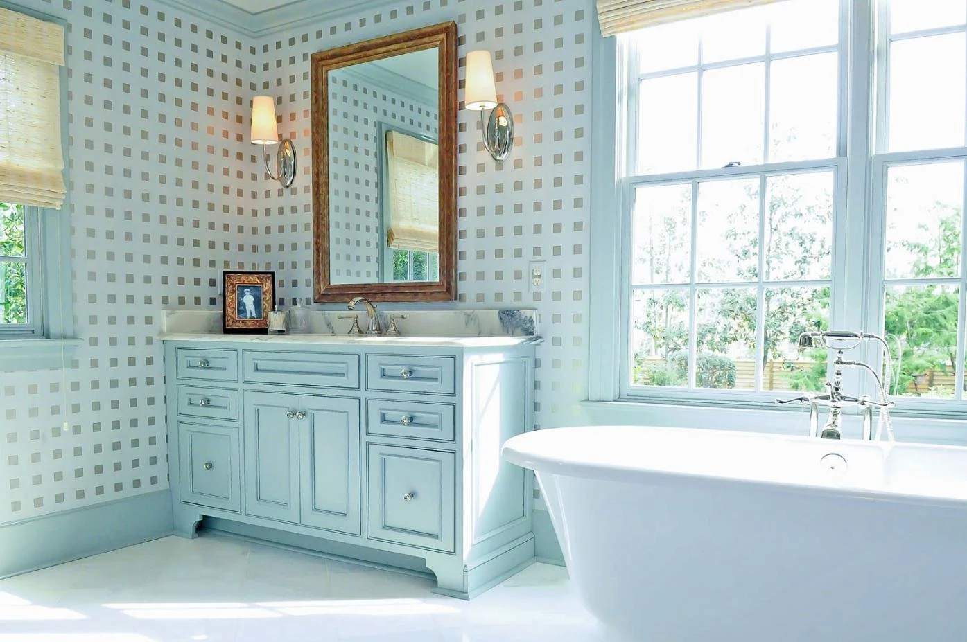 Bright traditional bathroom with a white vanity, patterned wallpaper, large windows, and a freestanding soaking tub.
