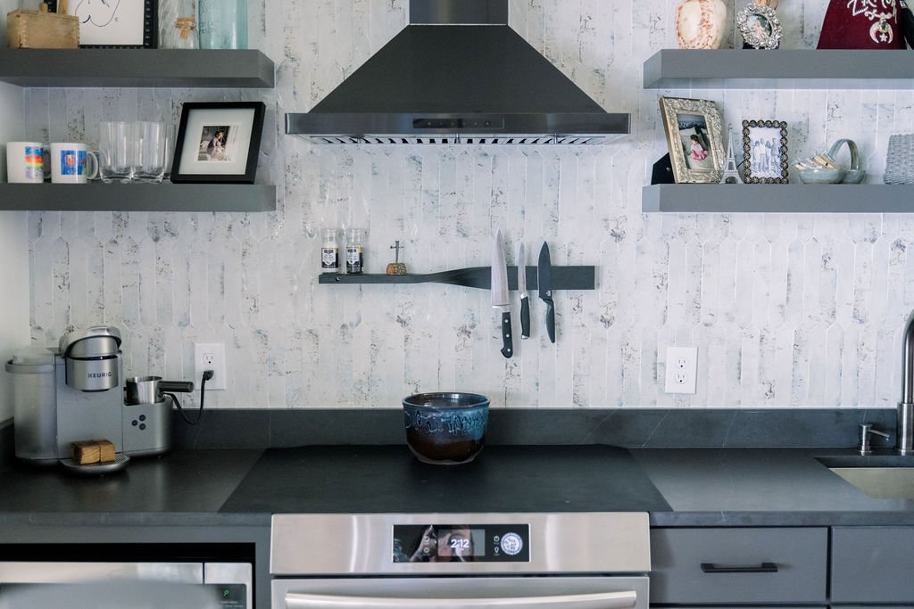 Textured white tile backsplash with dark floating shelves, black range hood and dark kitchen countertops