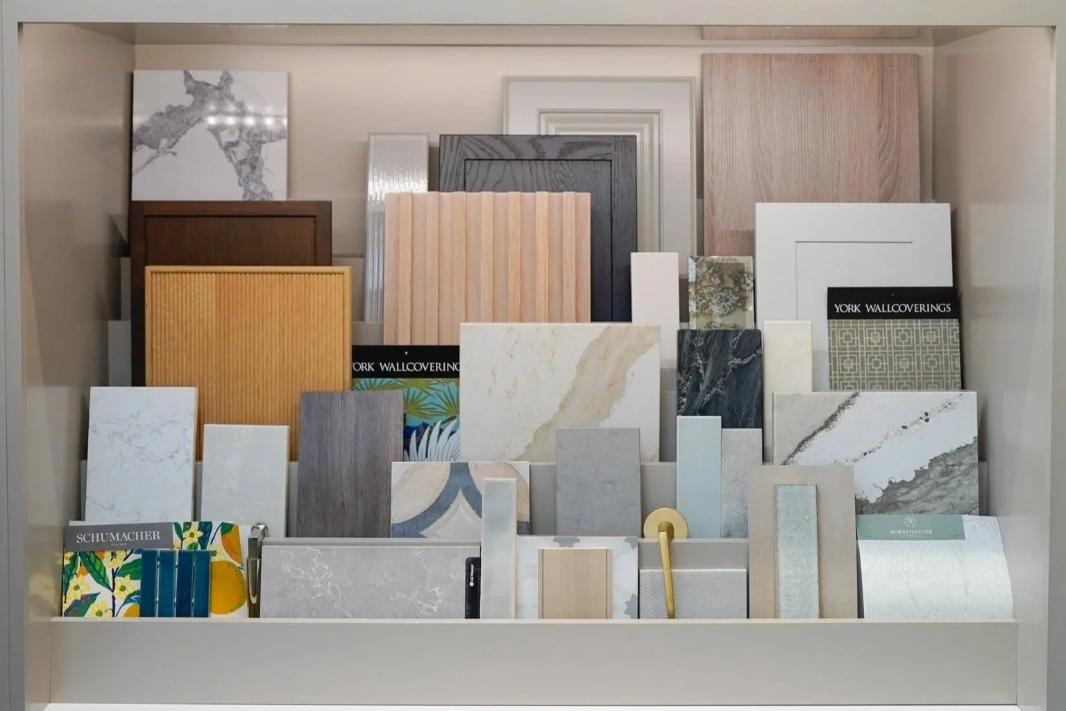 Display of custom cabinet door samples in various styles and finishes, including reeded wood panels, painted inset doors, slab fronts, and textured designs, arranged alongside countertop, tile, and wallcovering samples in a design showroom.