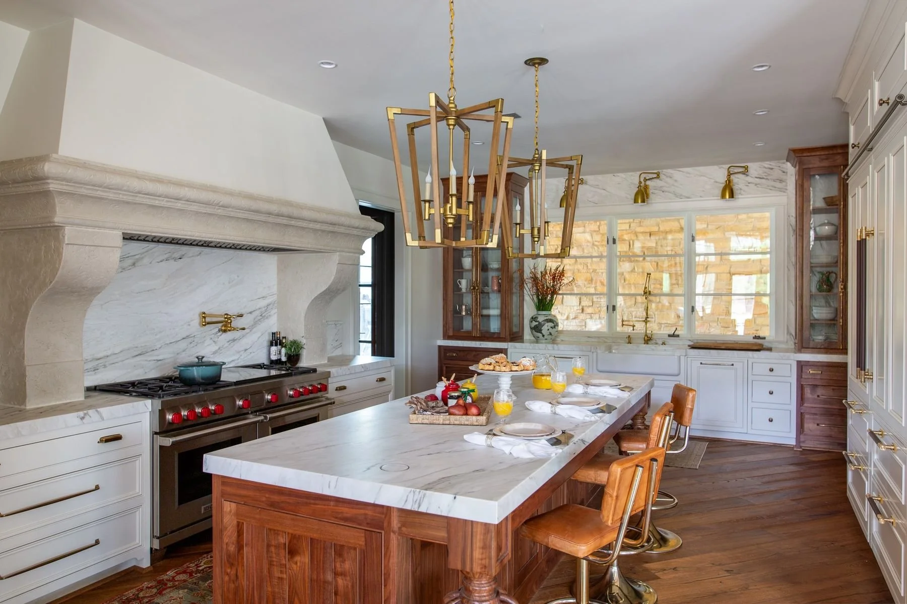 Luxury kitchen featuring a walnut island with marble countertop, custom white cabinetry, brass lighting, stone range hood, marble backsplash, and high-end appliances in an Alabama home.