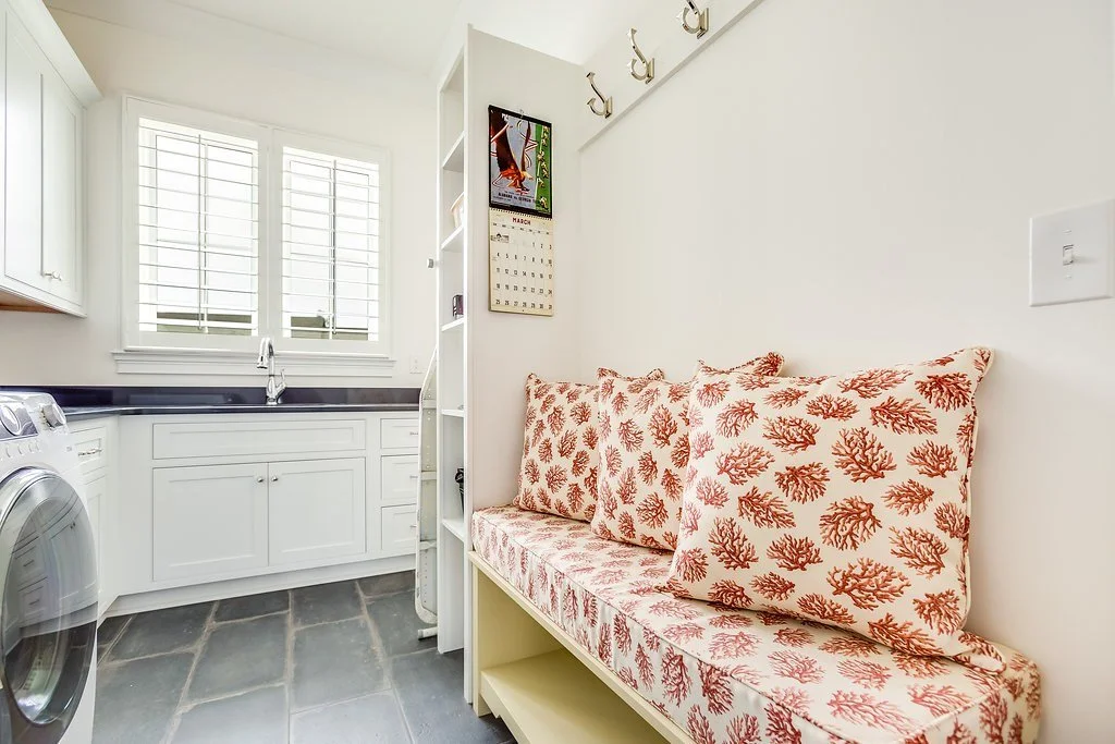 Bright laundry room with white cabinetry, sink, front-loading washer and built-in bench with patterned cushions and wall hooks.