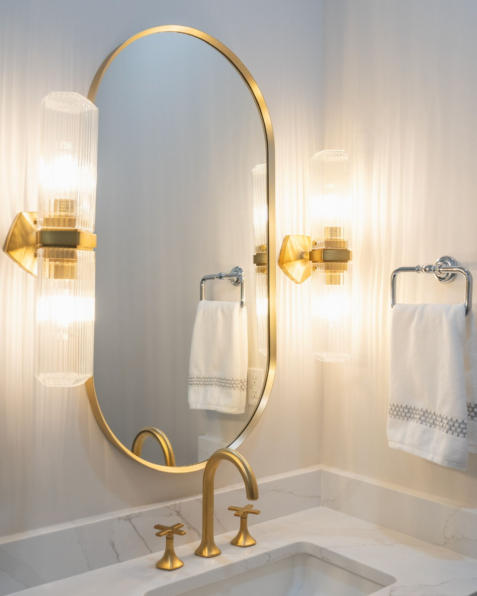 Close-up of fluted glass bathroom sconces mounted beside an arched brass-framed mirror above a quartz vanity countertop.