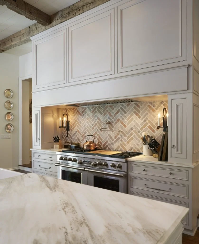 Detailed cabinetry frames a professional range, integrated hood, and herringbone backsplash to create an elegant kitchen focal point.