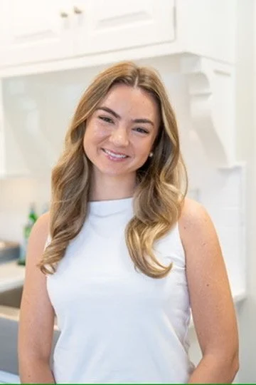 A young woman with long, wavy blonde hair smiling and wearing a navy blue dress with embroidered sleeves, standing in front of a light-colored wall with a blue palm leaf pattern.