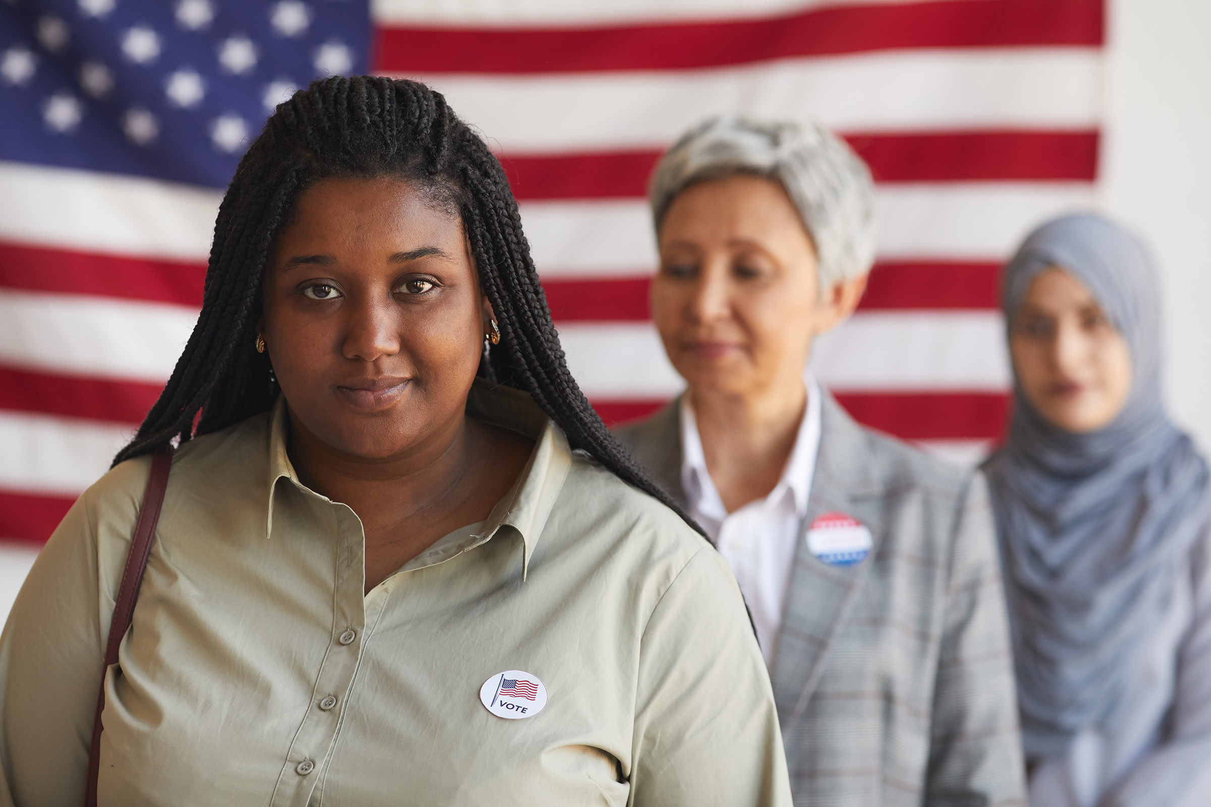 Three diverse women are standing in front of the American flag, waiting to vote.