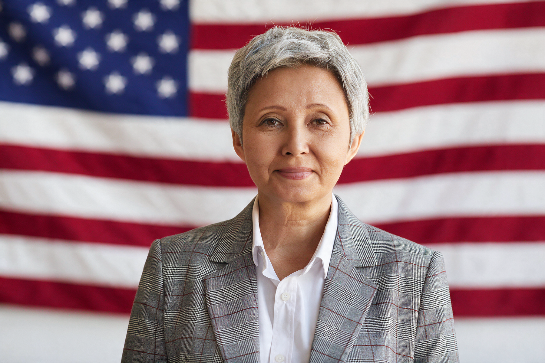 An older woman in a gray blazer standing in front of the American flag.