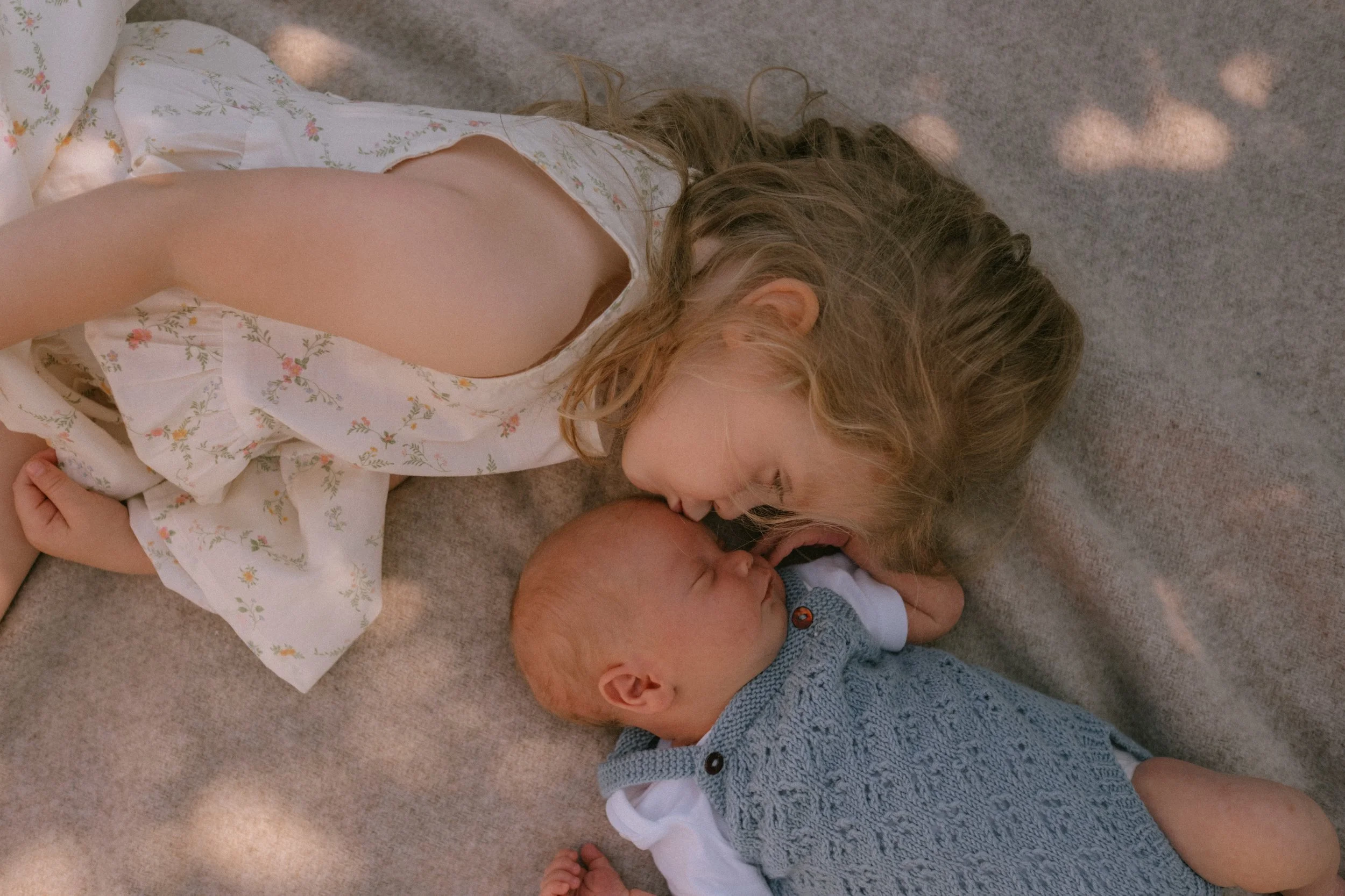 En baby i blå klær ligger på et pled i en hage, en jente kysser babyen på pannen. Søsken. Hage, familiefotografering.