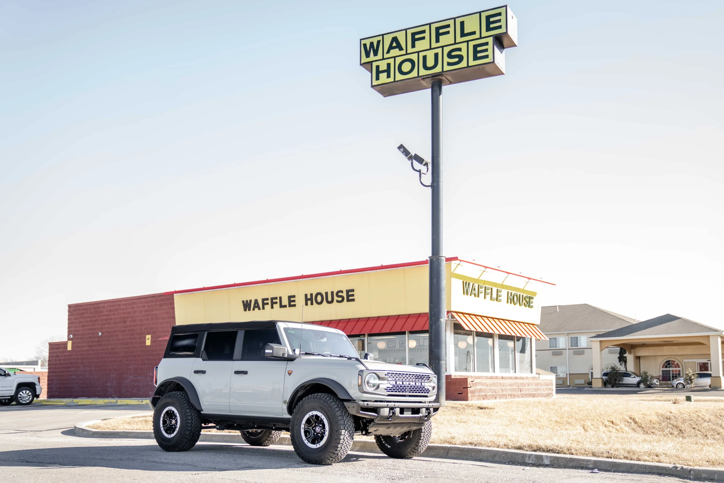 A white Ford Bronco parked in front of a Waffle House restaurant under a clear sky.