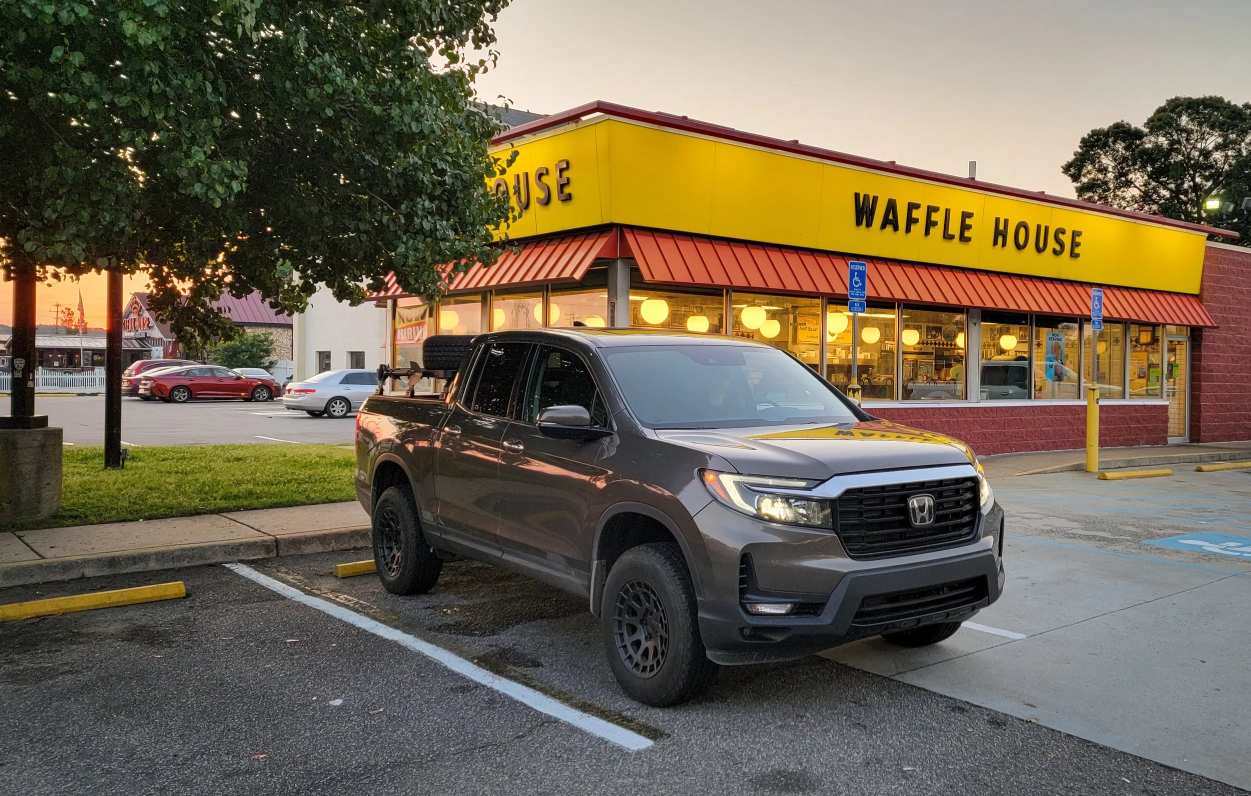 A Waffle House restaurant with a bright yellow sign and red roof trim, located in a parking lot. Several cars are parked, including a gray Honda Ridgeline in the foreground, and there are trees and other buildings nearby. The scene appears to be at s