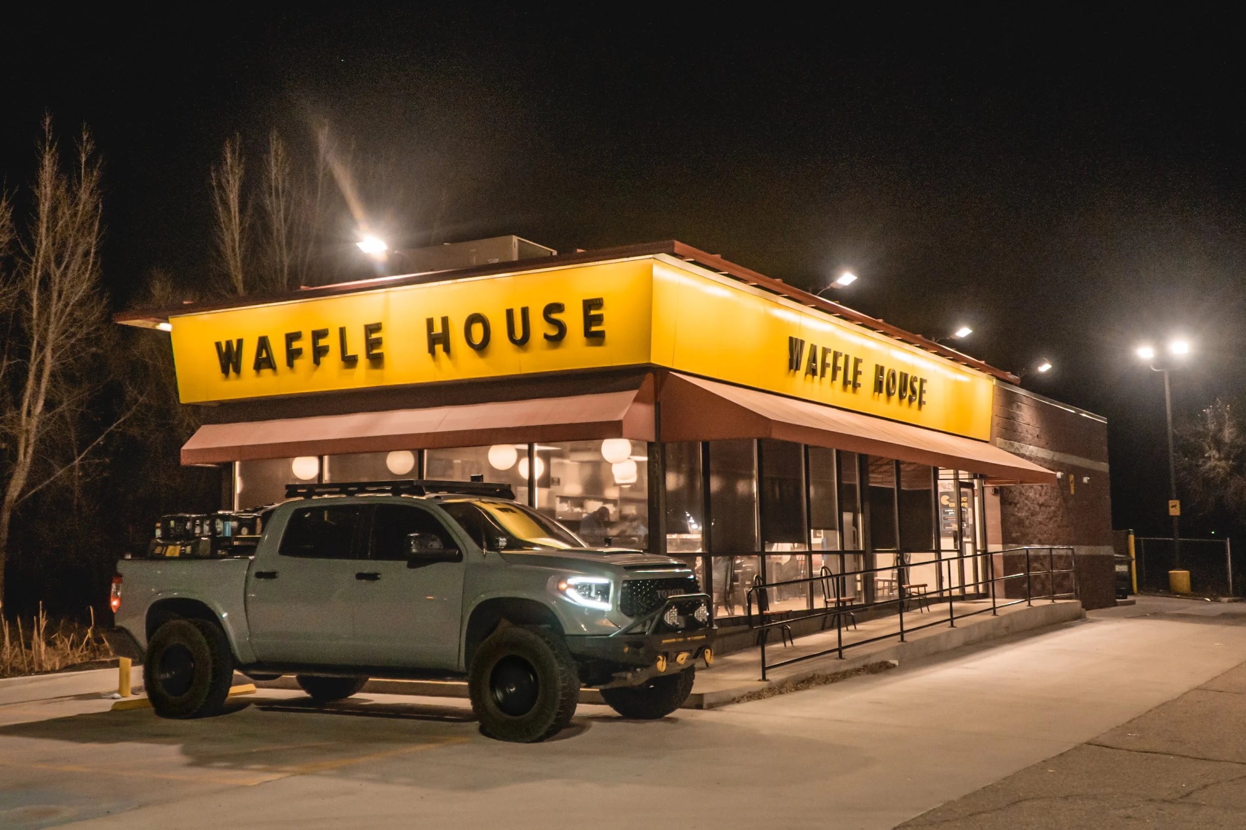 Nighttime view of a Waffle House restaurant with a bright yellow sign and parked truck in front, illuminated by exterior lights. Trees are visible in the background against the dark sky.