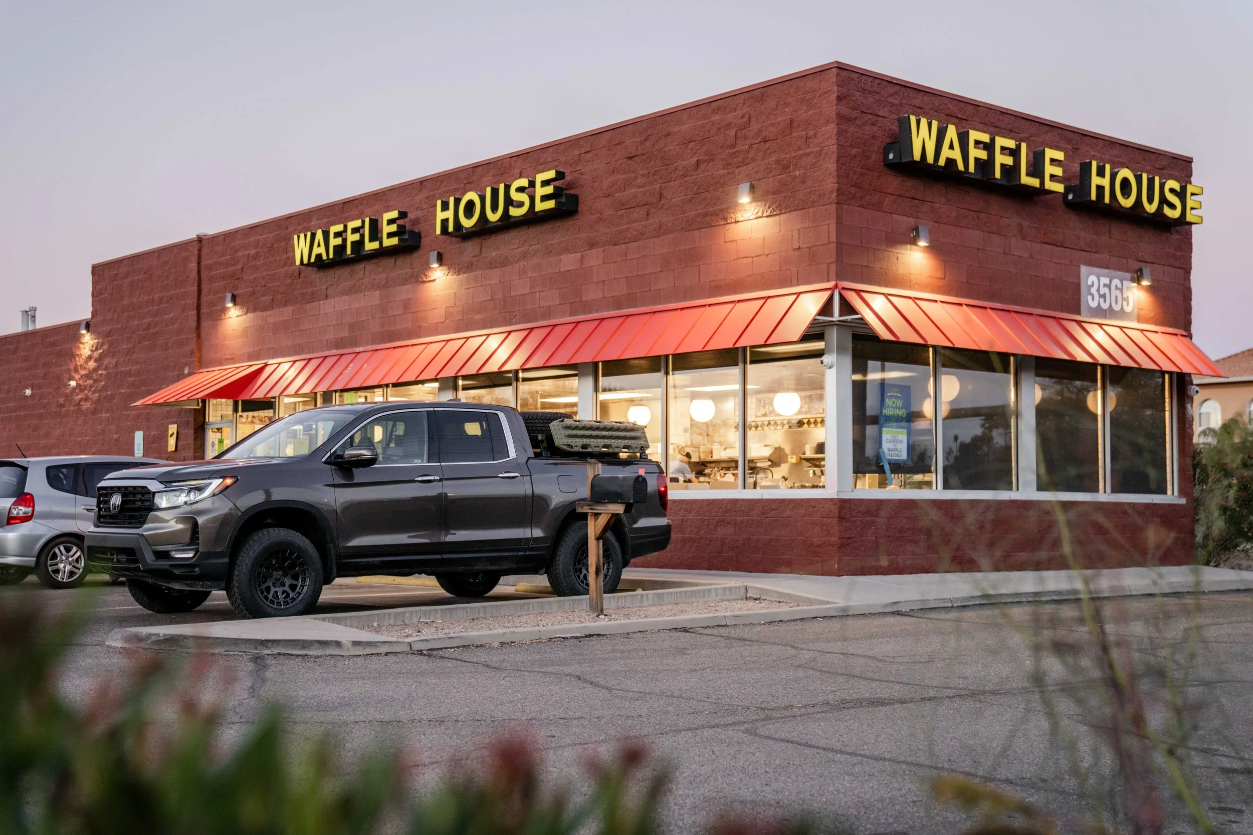 A fast food restaurant called 'Waffle House' with red brick exterior, yellow sign, and red awning, parked cars in front, and a street view at dusk.